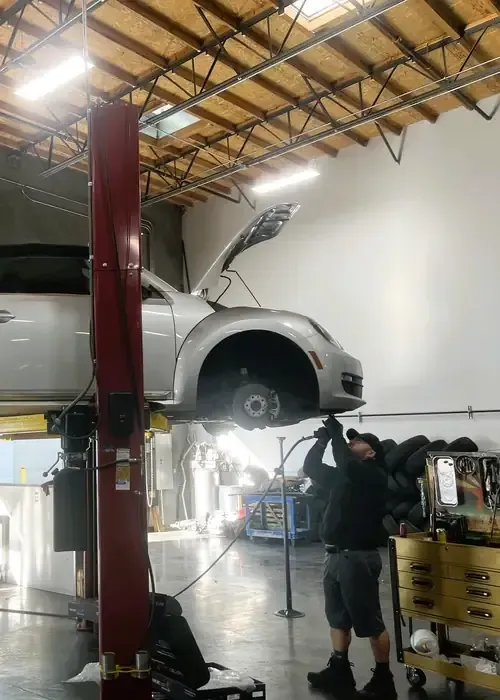 A mechanic in an auto repair shop works on a silver car lifted on a hydraulic hoist with its hood open.