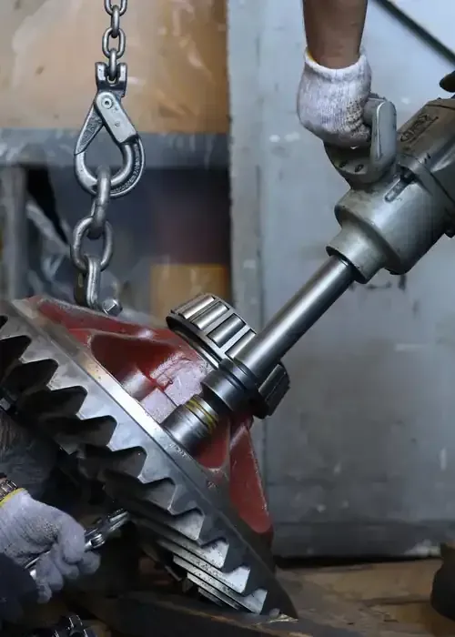 A worker uses a power tool and wrench to maintain a large metal gear assembly suspended by a chain in a workshop.