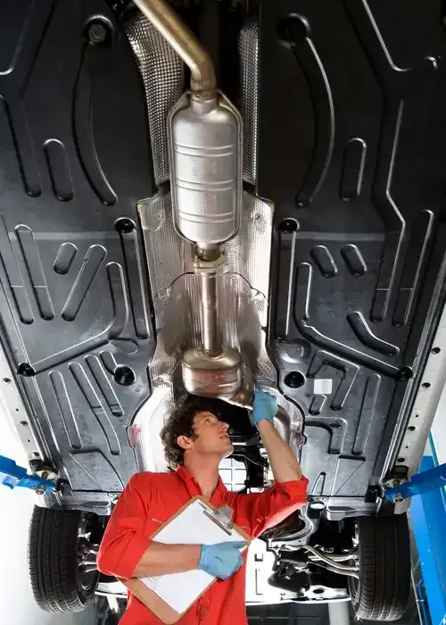 A mechanic in a red uniform and blue gloves inspects the underside of a car raised on a lift, holding a clipboard.