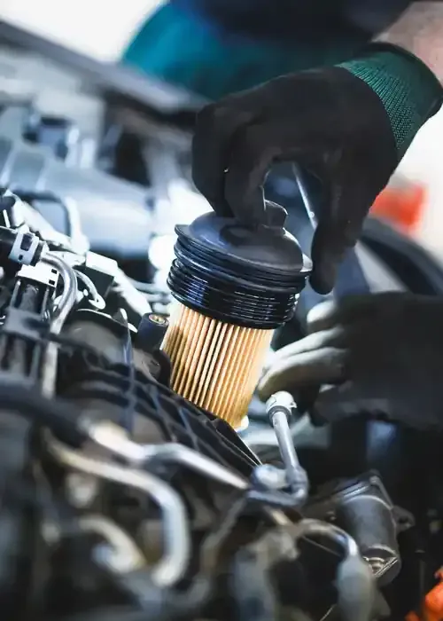 A mechanic in black gloves removes a cylindrical pleated oil filter from a vehicle engine bay.
