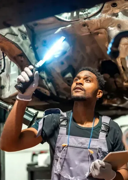 A mechanic wearing gray overalls holds a bright work light while inspecting the undercarriage of a vehicle in a garage.