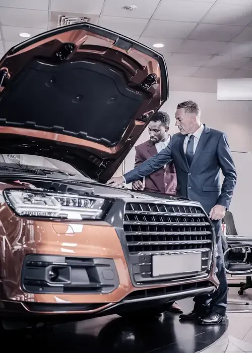 Two people in suits stand in a showroom, inspecting the open engine bay of a metallic bronze car.