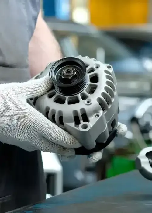 A person wearing work gloves holding a car alternator in a workshop setting.