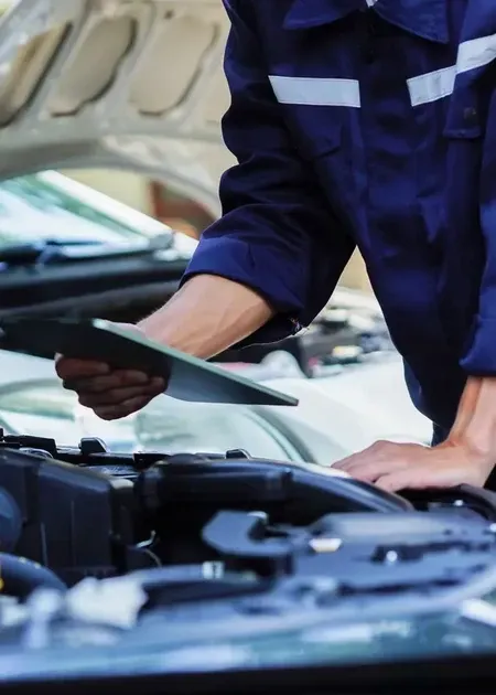 A mechanic in a blue uniform examines a car engine with the hood open, holding a clipboard.