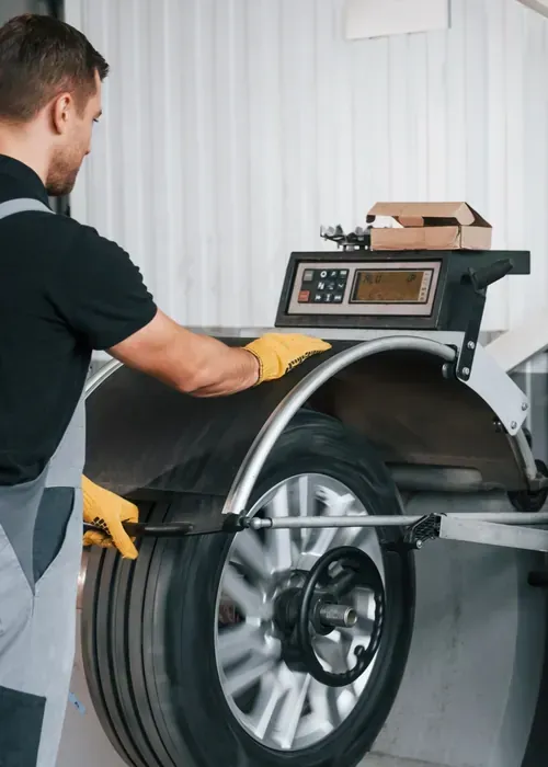 A technician in a uniform and yellow gloves uses a tire balancing machine to inspect a car wheel in a workshop.