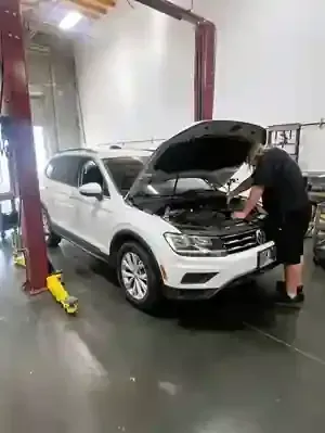 A mechanic works on the rear underside of a white Dodge Challenger lifted on a hydraulic hoist in an auto shop.