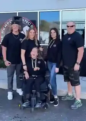 Five people posing outdoors in front of a building; one person is seated in a power wheelchair, others stand behind them.