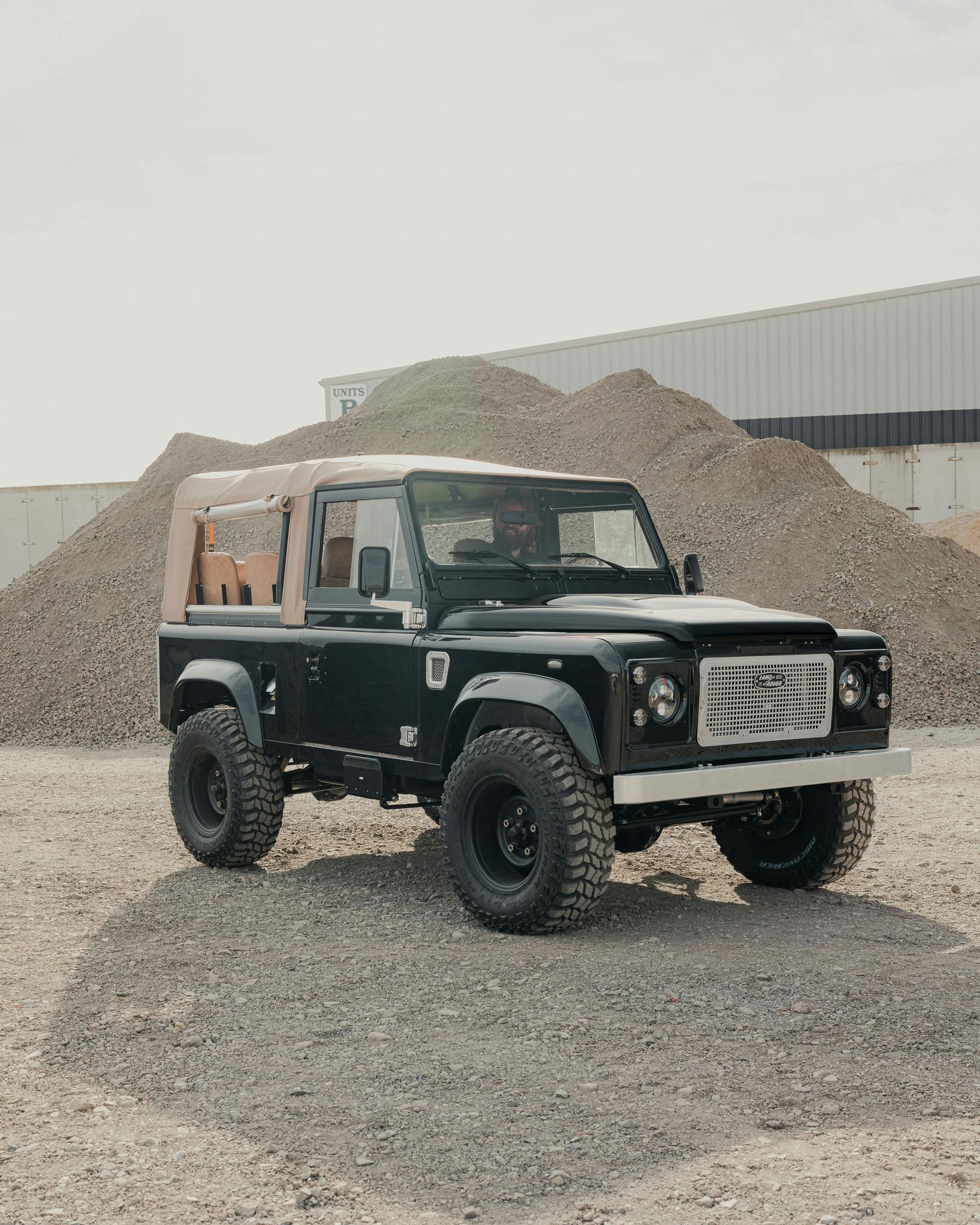 A black Land Rover Defender with a tan soft top parked on a gravel lot in front of large piles of earth.