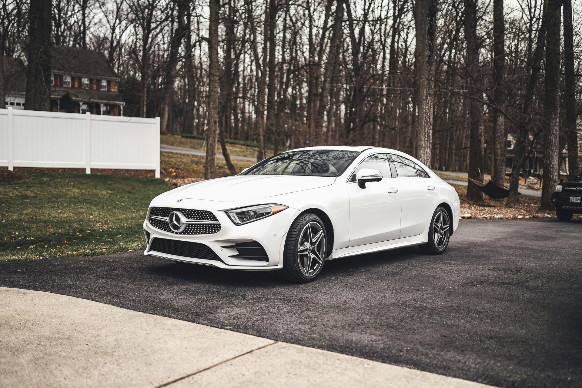 A white Mercedes-Benz CLS sedan parked on an asphalt driveway in front of a house and trees.