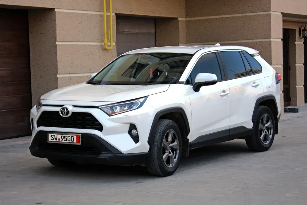 A white Toyota RAV4 SUV parked on a paved driveway in front of a building with beige walls and garage doors.