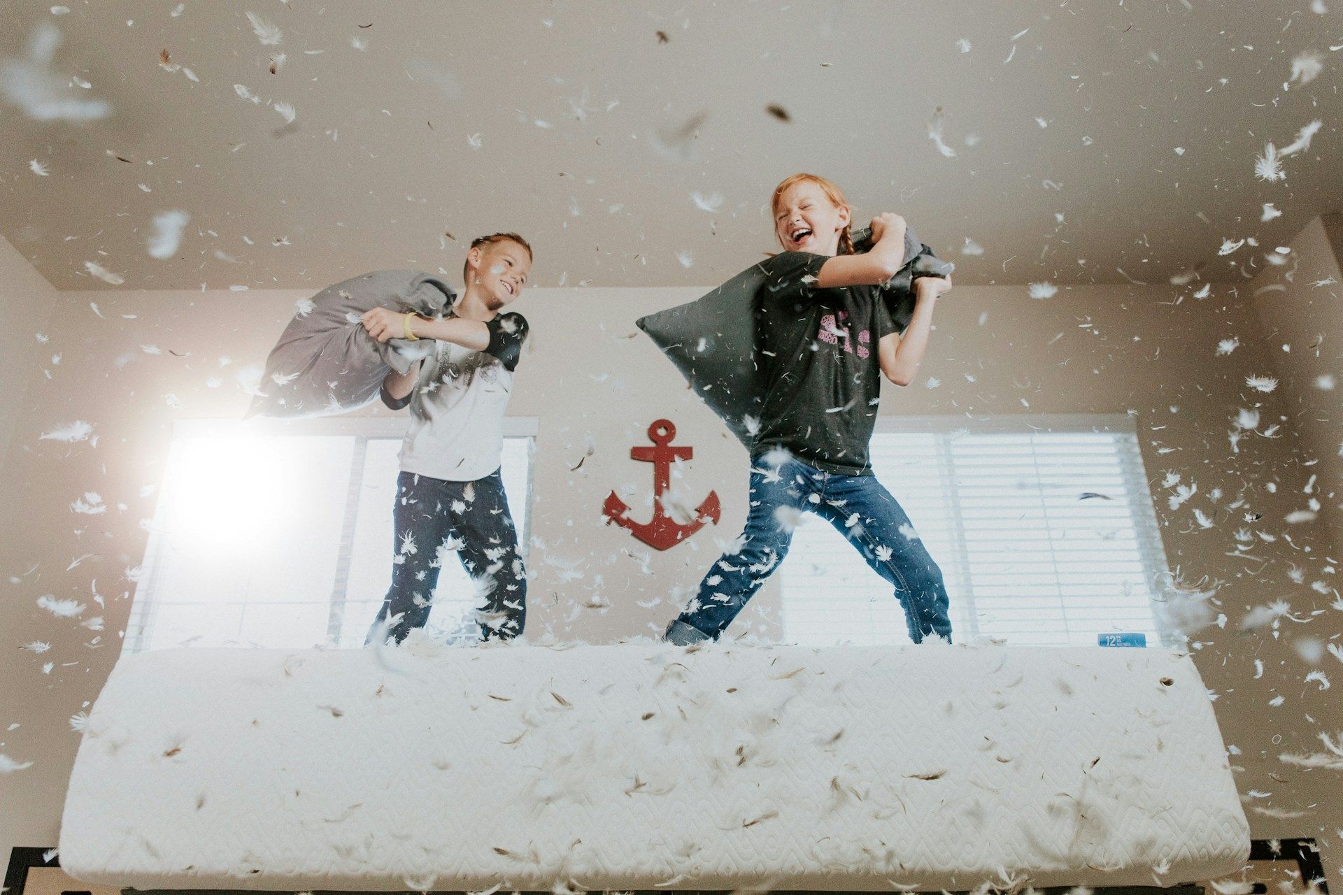Two boys in a pillow fight on a bed. Feathers fly in the air; one laughs, one swings a pillow.