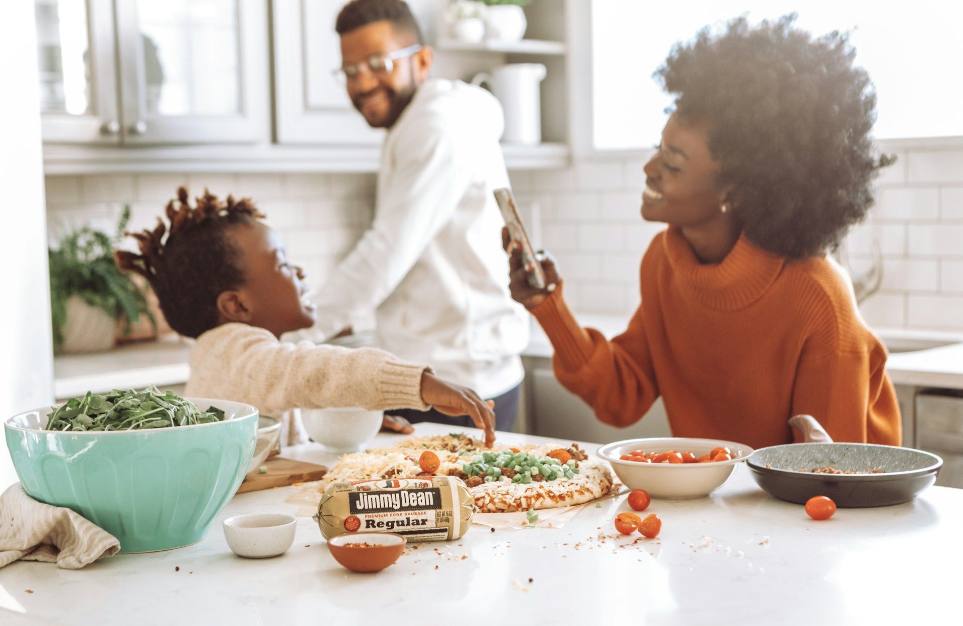 Family in kitchen preparing food: woman smiling, child reaching, man in background, pizza and ingredients on counter.
