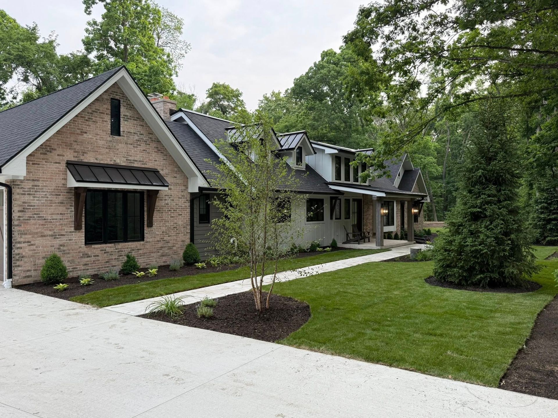 Suburban brick house with black awnings, white driveway, and manicured lawn surrounded by trees