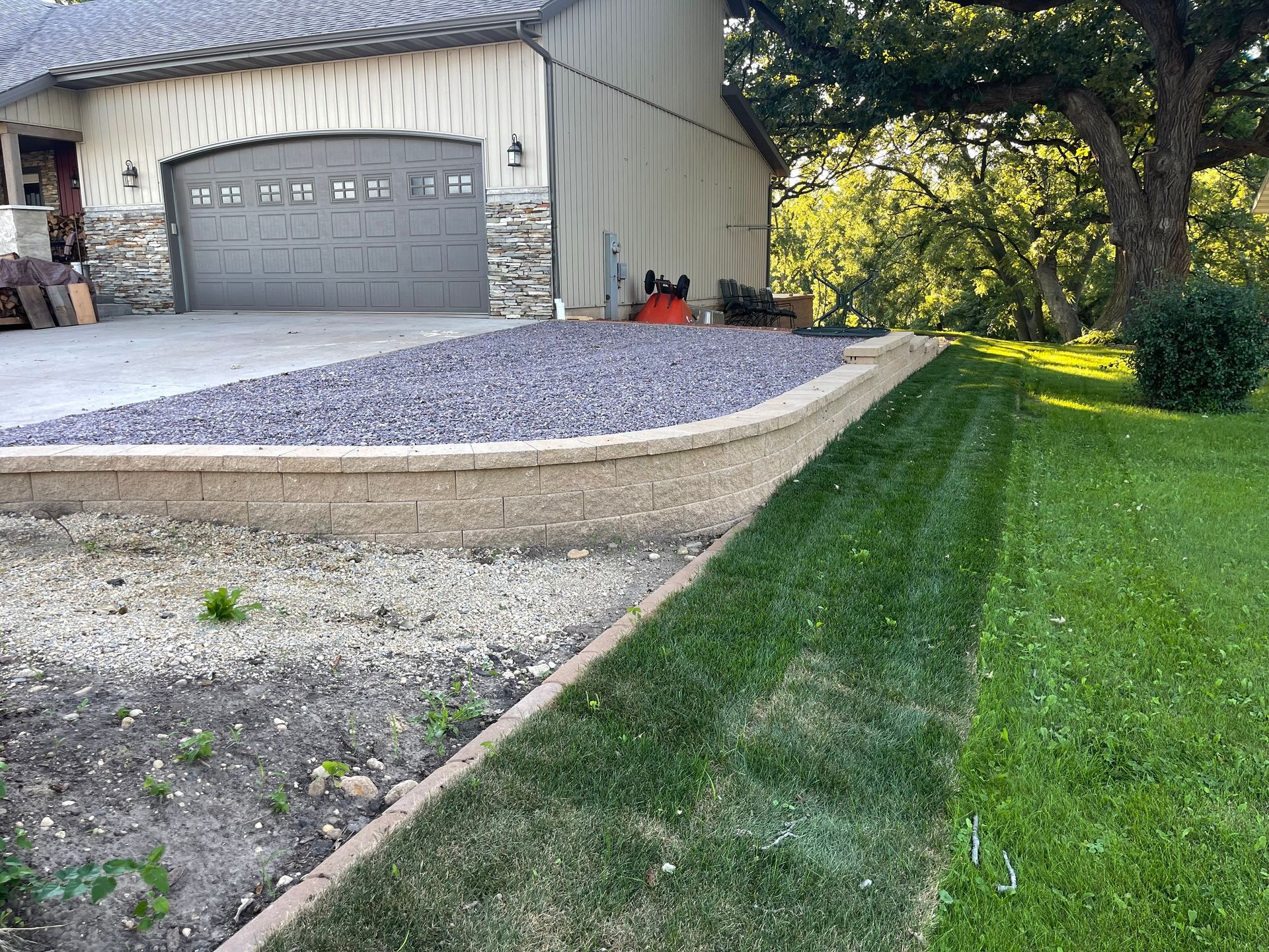 Garage with gravel driveway beside a sloped green lawn and a tree-shaded yard