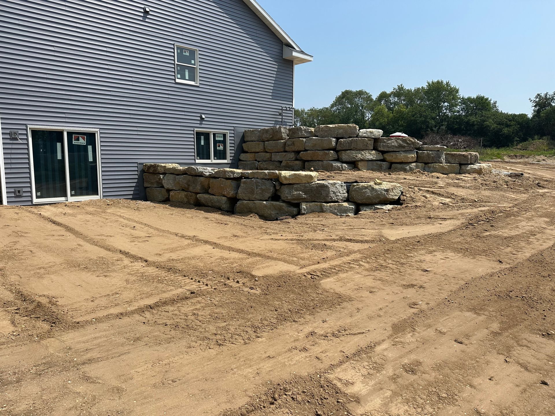 Construction site with stacked stone blocks beside a gray house on sandy ground
