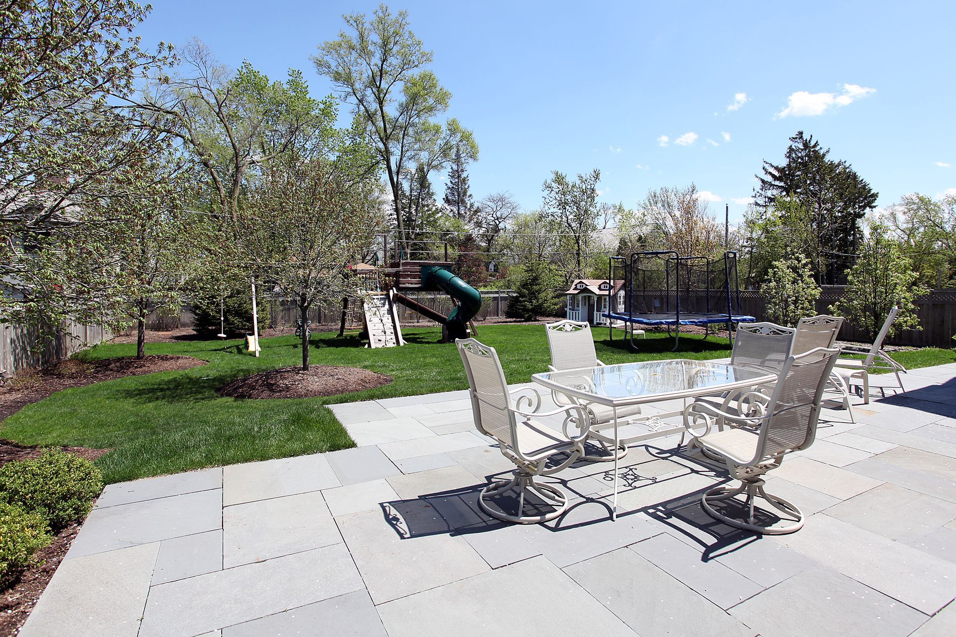 Patio seating area with white chairs and table beside a grassy park and playground on a sunny day