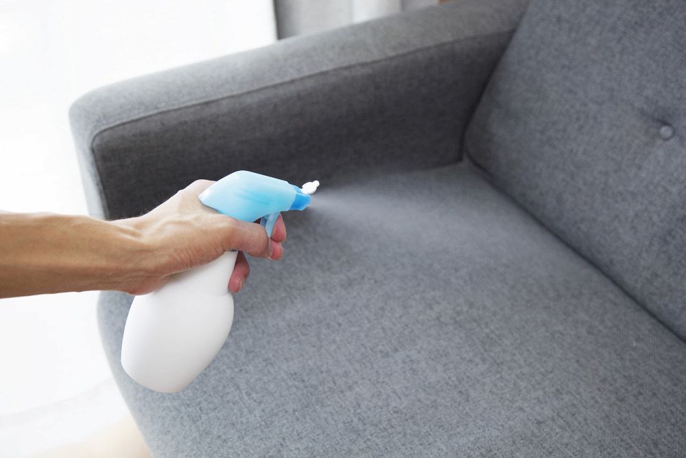 Person spraying upholstery with a blue and white spray bottle. Gray sofa in the background. — EcoAir Airconditioning in Humpty Doo, NT
