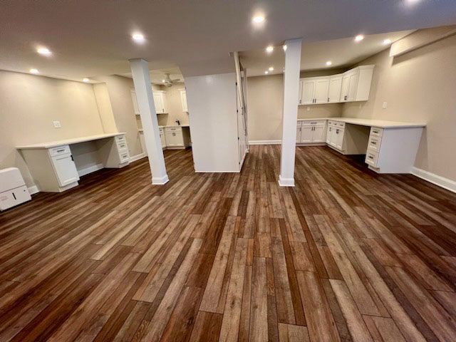 Finished basement with wood-look flooring, white desks and cabinets, recessed lighting, and support columns.