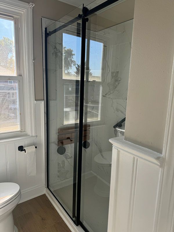 Bathroom with glass shower door, black frame, white wainscoting, and toilet near a window.