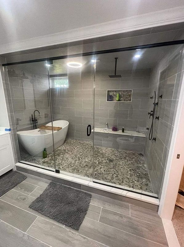 Luxurious bathroom with a glass-enclosed shower, freestanding tub, pebble flooring, gray tiles, and multiple showerheads.