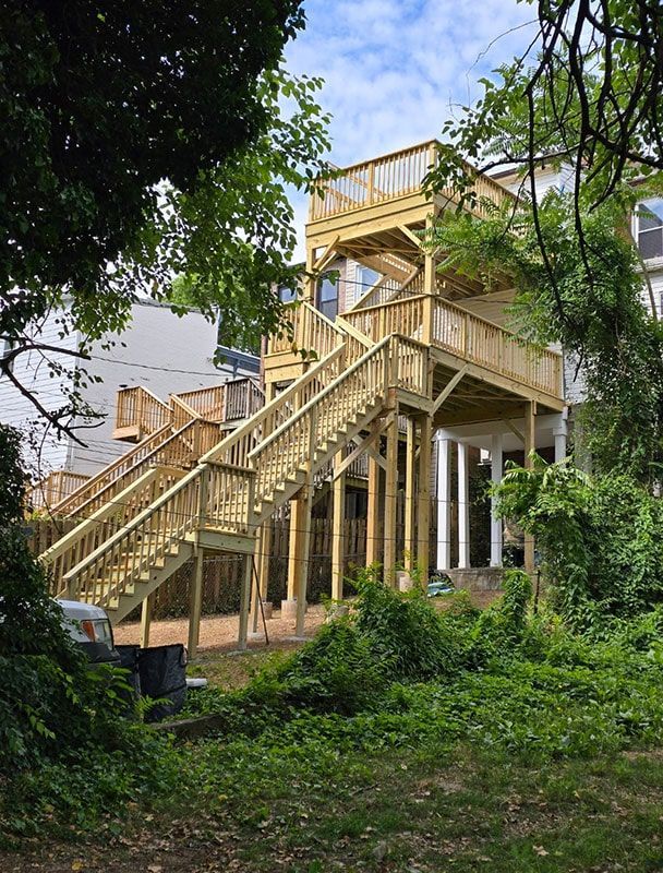 Multi-level wooden deck with stairs in a backyard setting, surrounded by greenery.