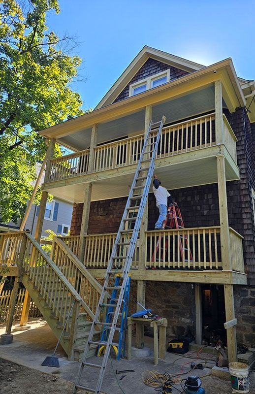 Two-story wooden deck addition to a house; two people on the upper deck, ladder extends to the top level.