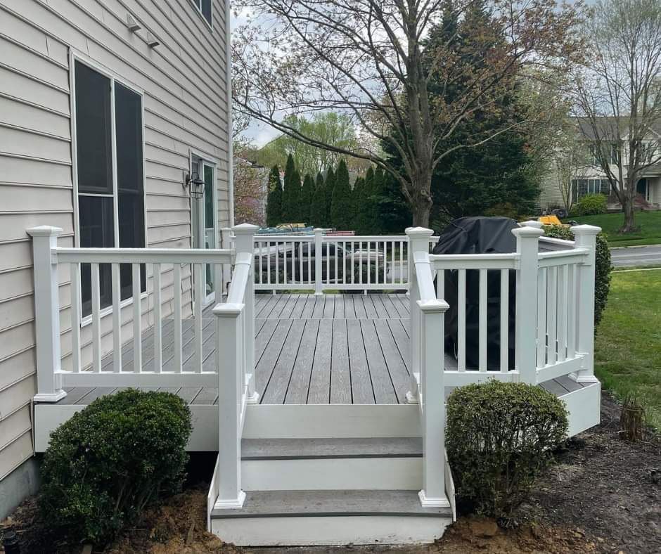 White deck with stairs, surrounded by a white railing. Shrubs in front. House with siding in background.