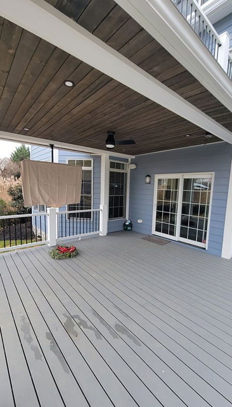 Covered deck with gray flooring, blue house, and wooden ceiling.  A sheer brown cloth hangs on the railing.