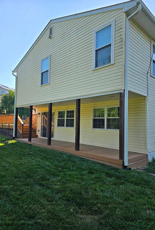 Back of a two-story yellow house with a covered wooden deck supported by dark-stained posts, green lawn in front.