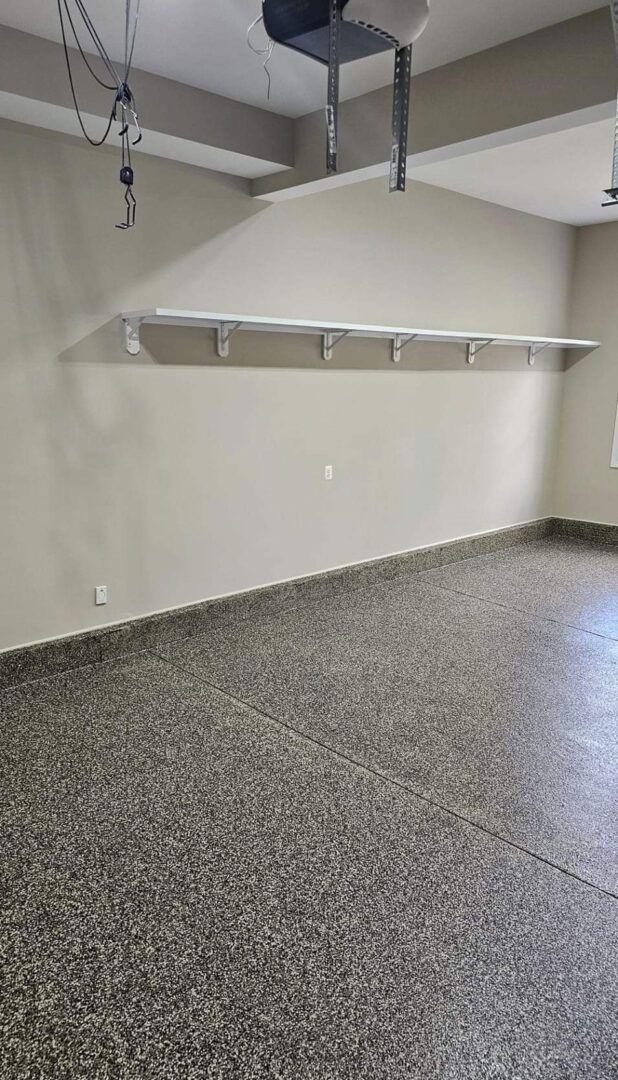 Empty garage interior with tan walls, white shelf, and dark speckled floor.