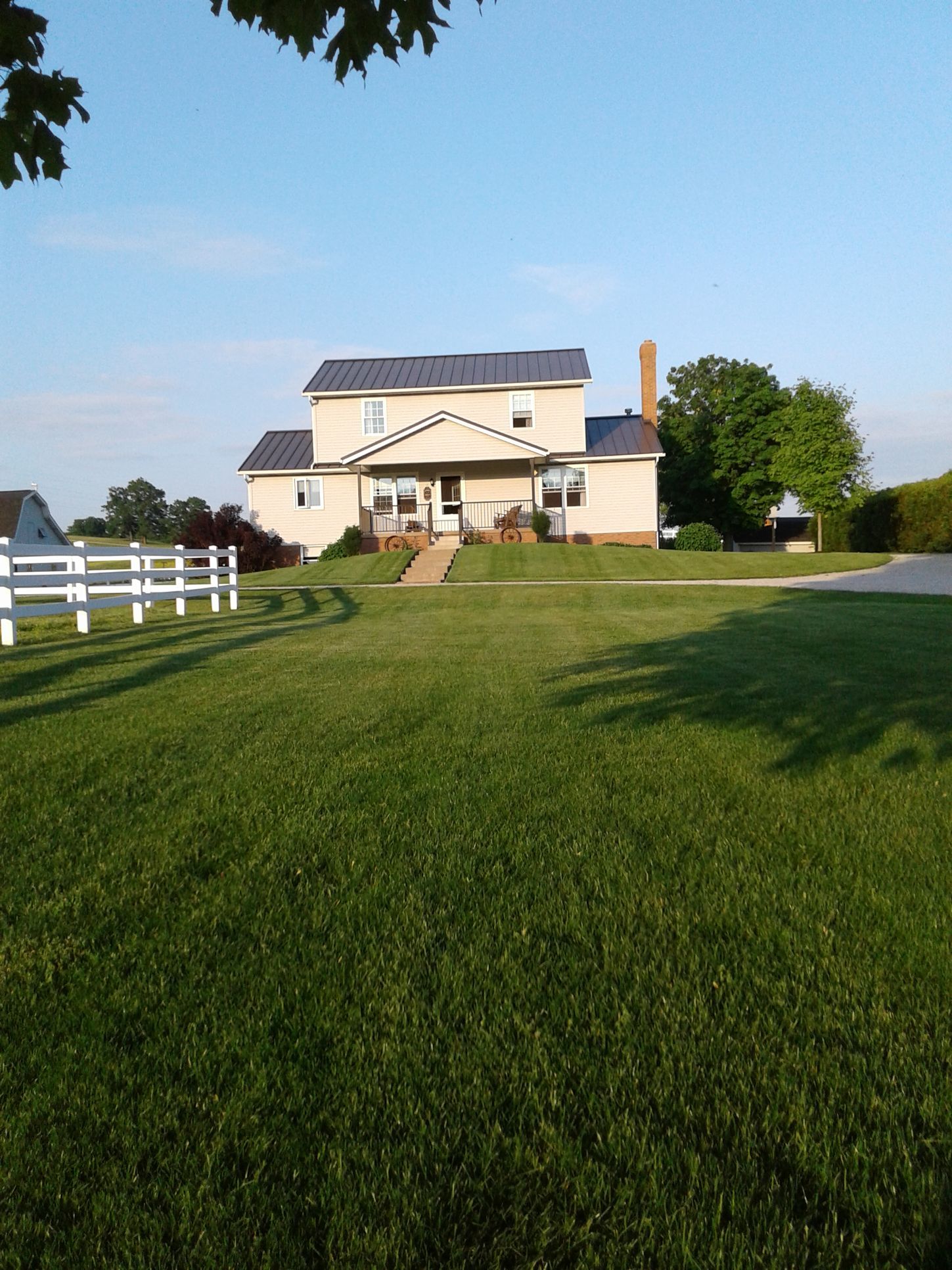 Two-story beige house with a gray roof, chimney, and porch. White fence and green lawn under a blue sky.