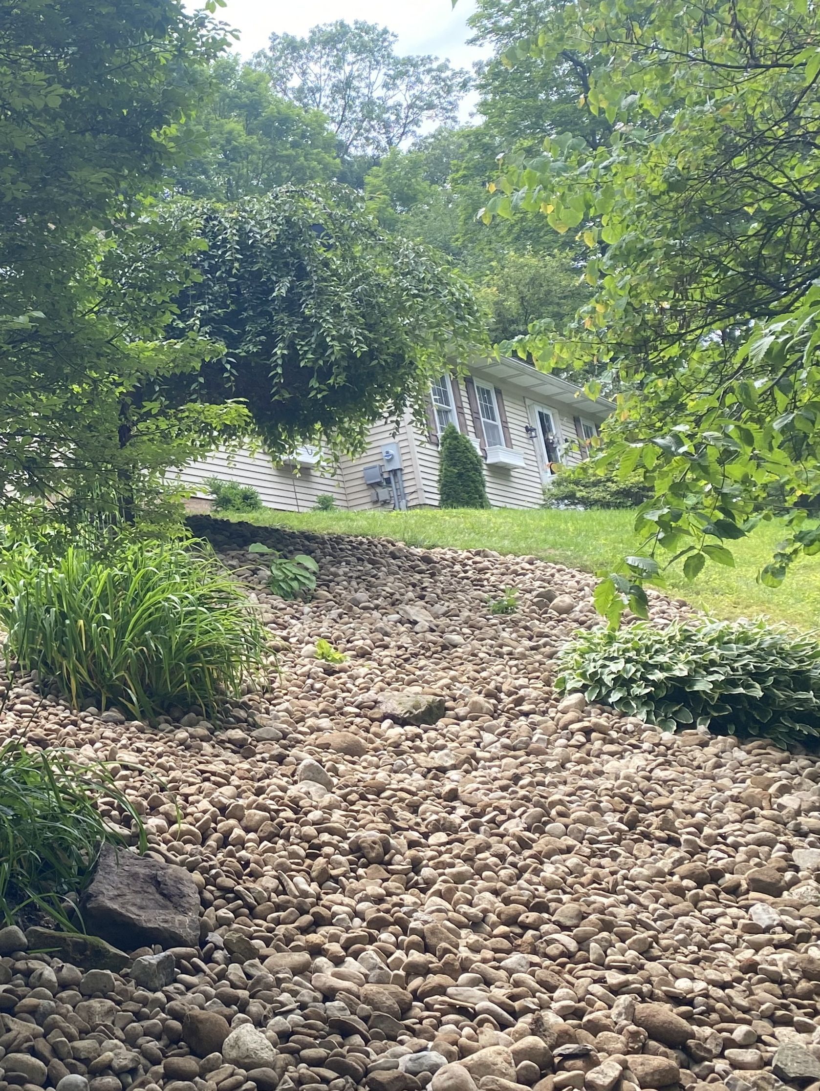 Rocky stream bed leading to a white house with trees in the background, sunny day.
