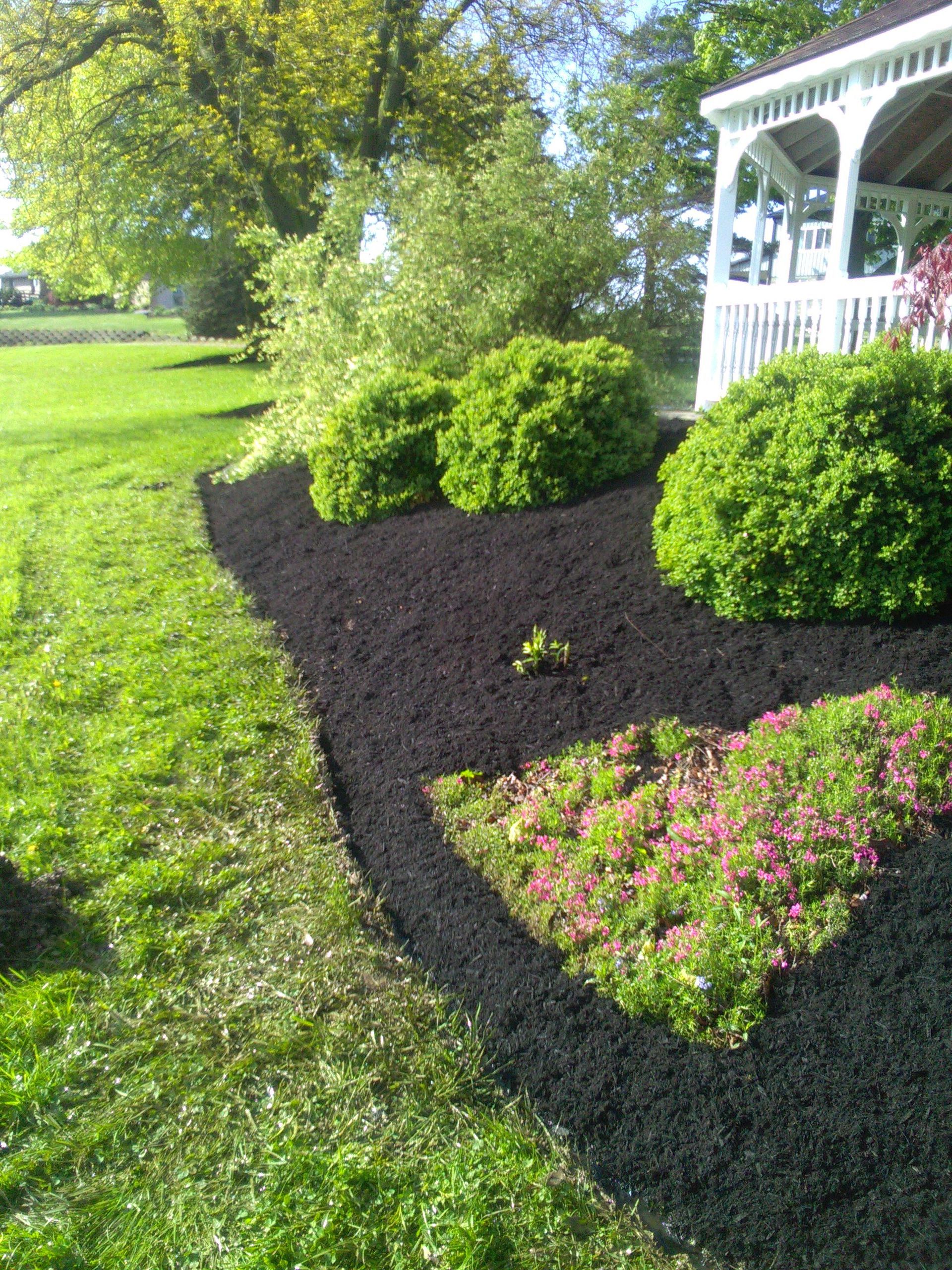 Green lawn with flowerbeds edged in black mulch, near a white gazebo.