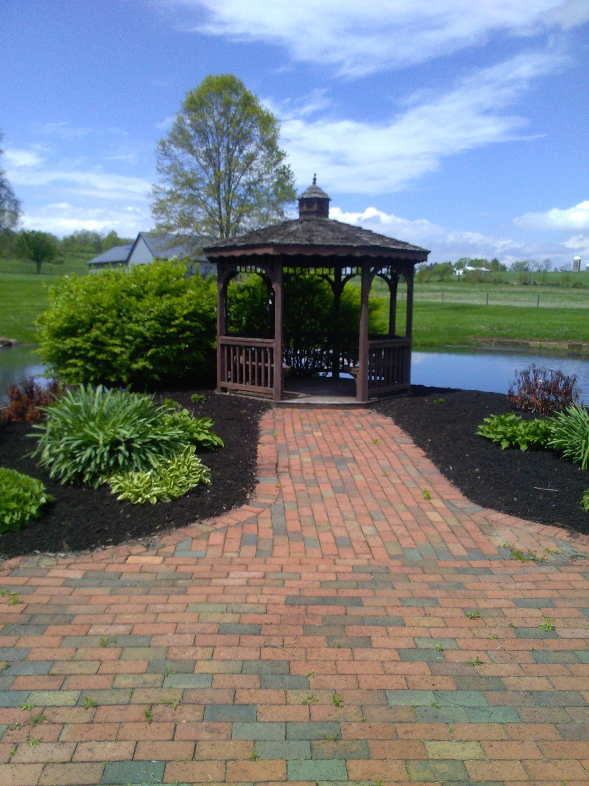 Gazebo with brick pathway near a pond, surrounded by landscaping, under a blue sky.