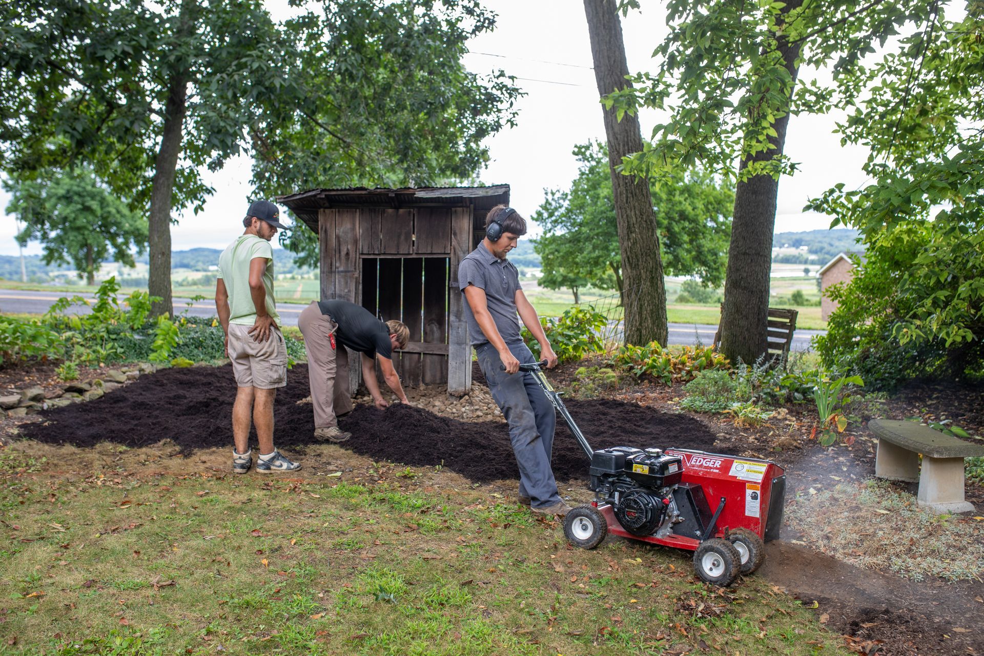 Three people mulching a garden bed near a small wooden shed with a rototiller.