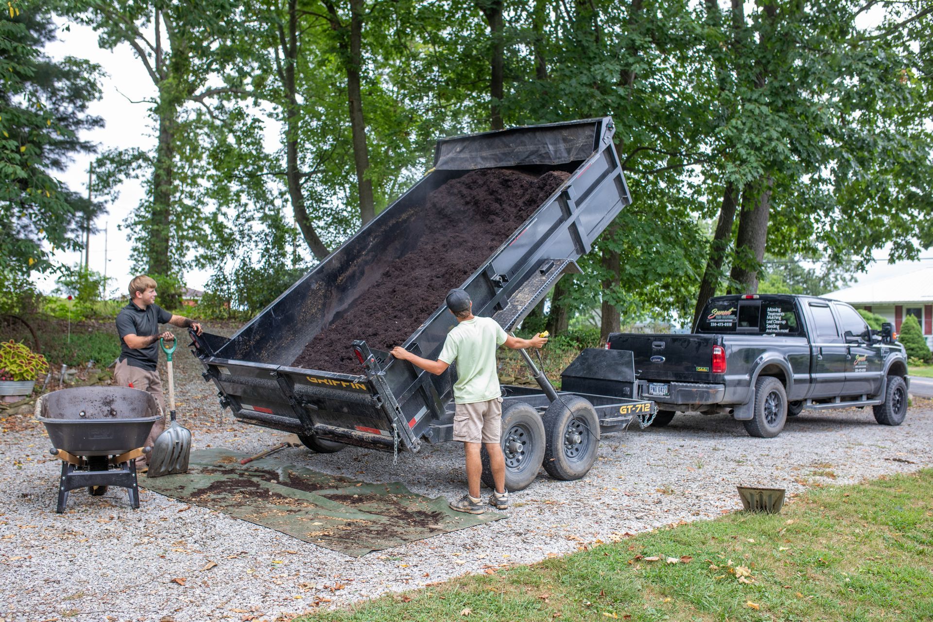 Two people unload mulch from a trailer hitched to a truck on a gravel driveway.