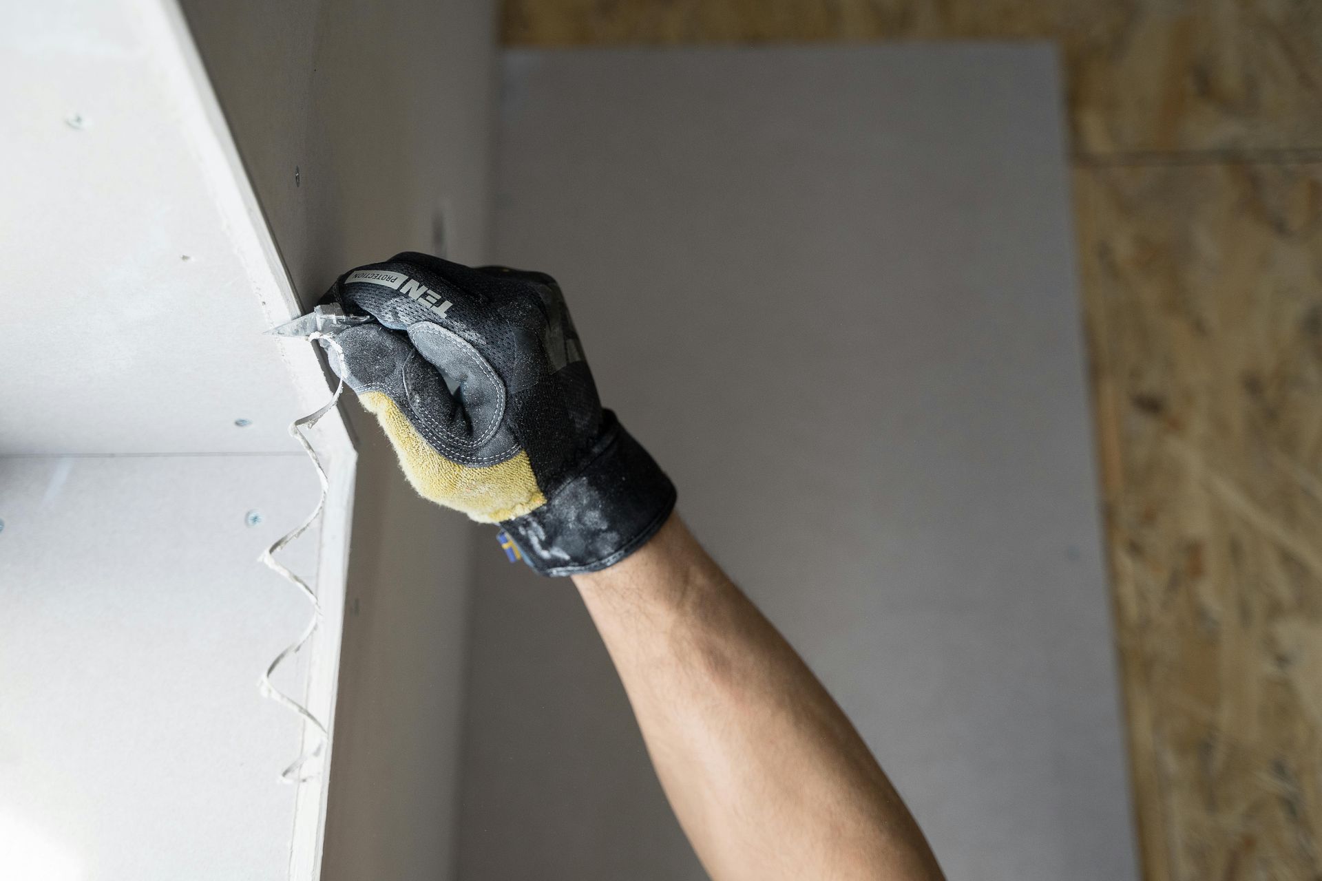 A gloved hand uses a utility knife to trim the edge of a white drywall panel installed in a building.