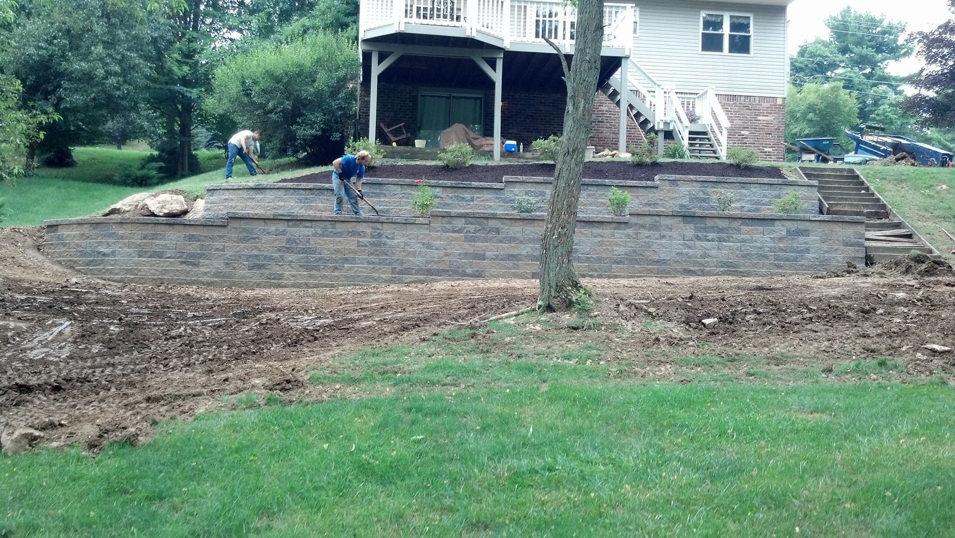 Workers constructing a tiered retaining wall near a house. Ground is bare; lawn in the foreground.