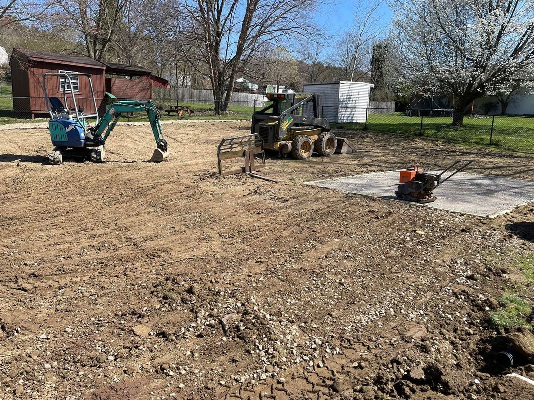 Construction site with mini excavator, skid steer, and compactor on bare earth; small shed in background.