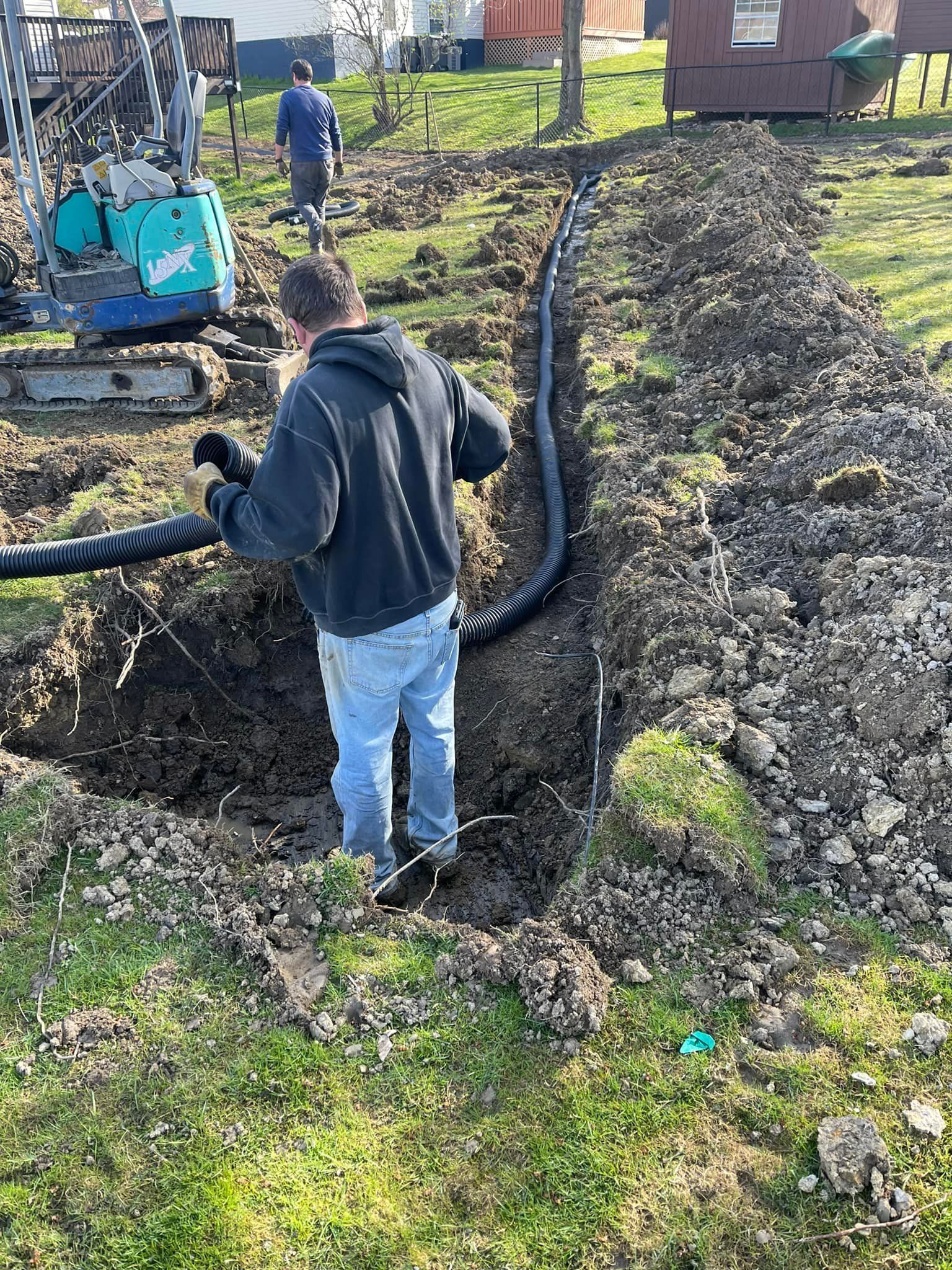 Man installing drainage pipe in a trench on a grassy lawn with construction equipment.