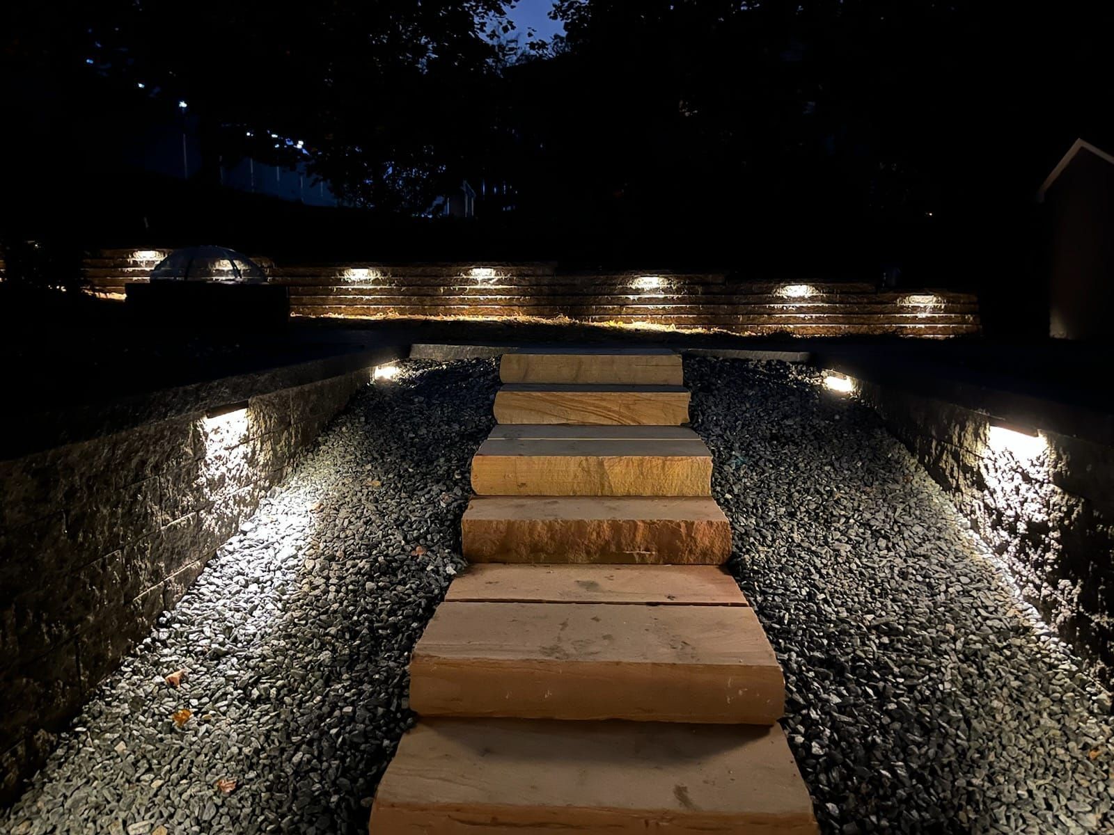 Stone steps illuminated by lights in a gravel pathway, leading to a wall with more lights at dusk.
