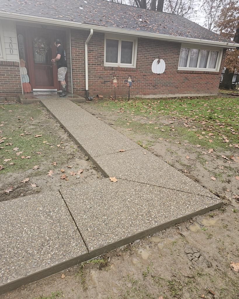 Person stands at a brick house entrance with a concrete path. Path is surrounded by mud and grass.