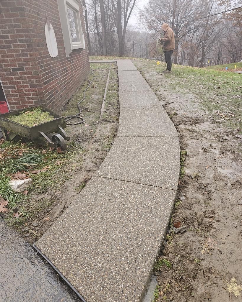 A concrete pathway curves past a brick house and muddy ground. A person stands at the top of the path.