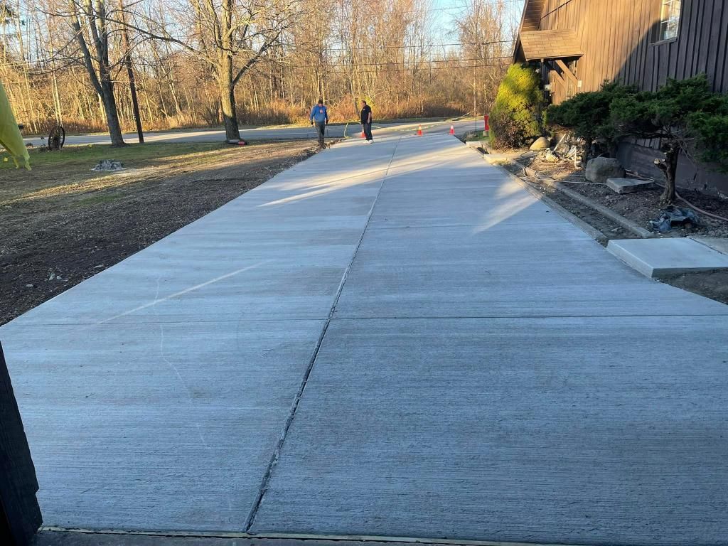 Long concrete driveway with two people on it, trees in the background, and a building on the right.