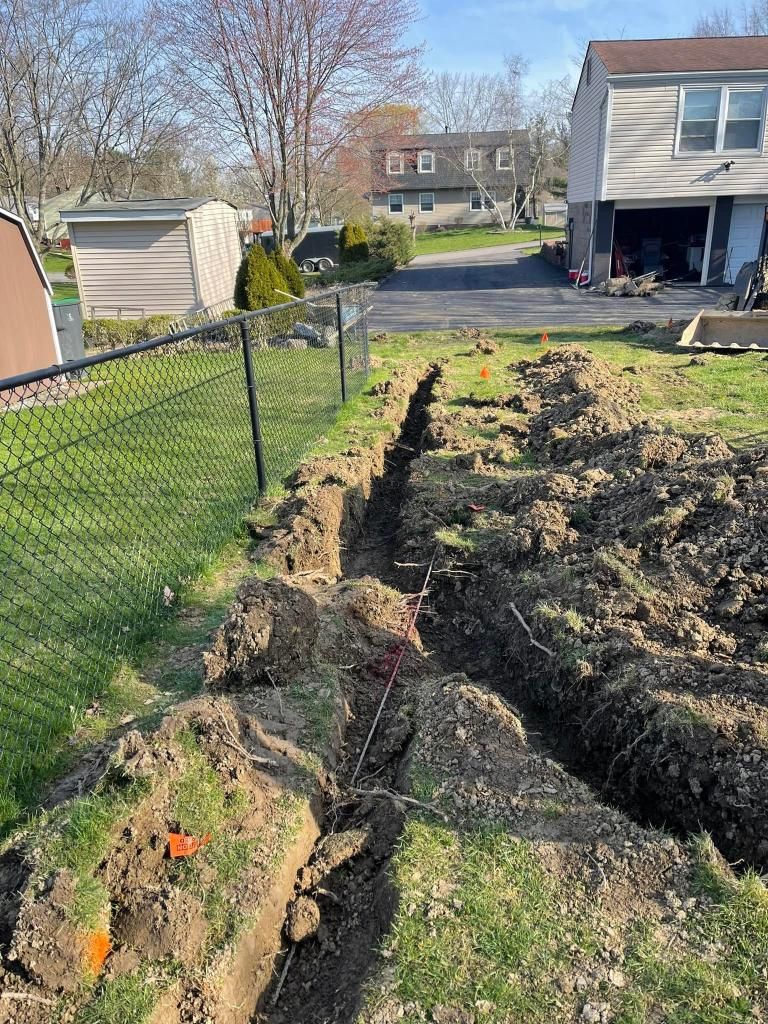 A trench dug in a backyard, alongside a black chain-link fence. Orange markers and dirt piles are present.