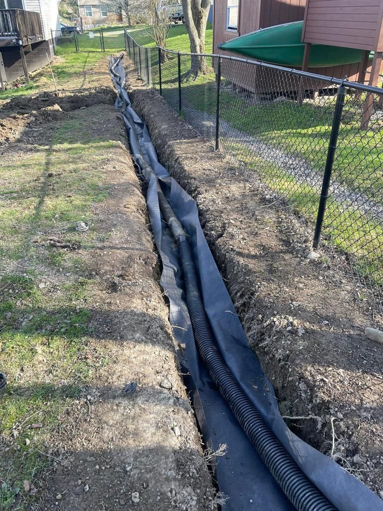 A trench in a yard with black fabric and corrugated pipe, next to a black fence.