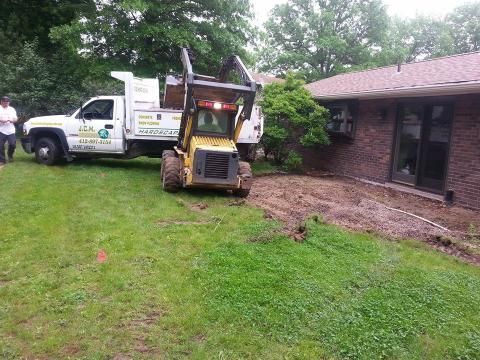 A yellow skid steer near a brown brick house, next to a white dump truck, in a grassy yard.