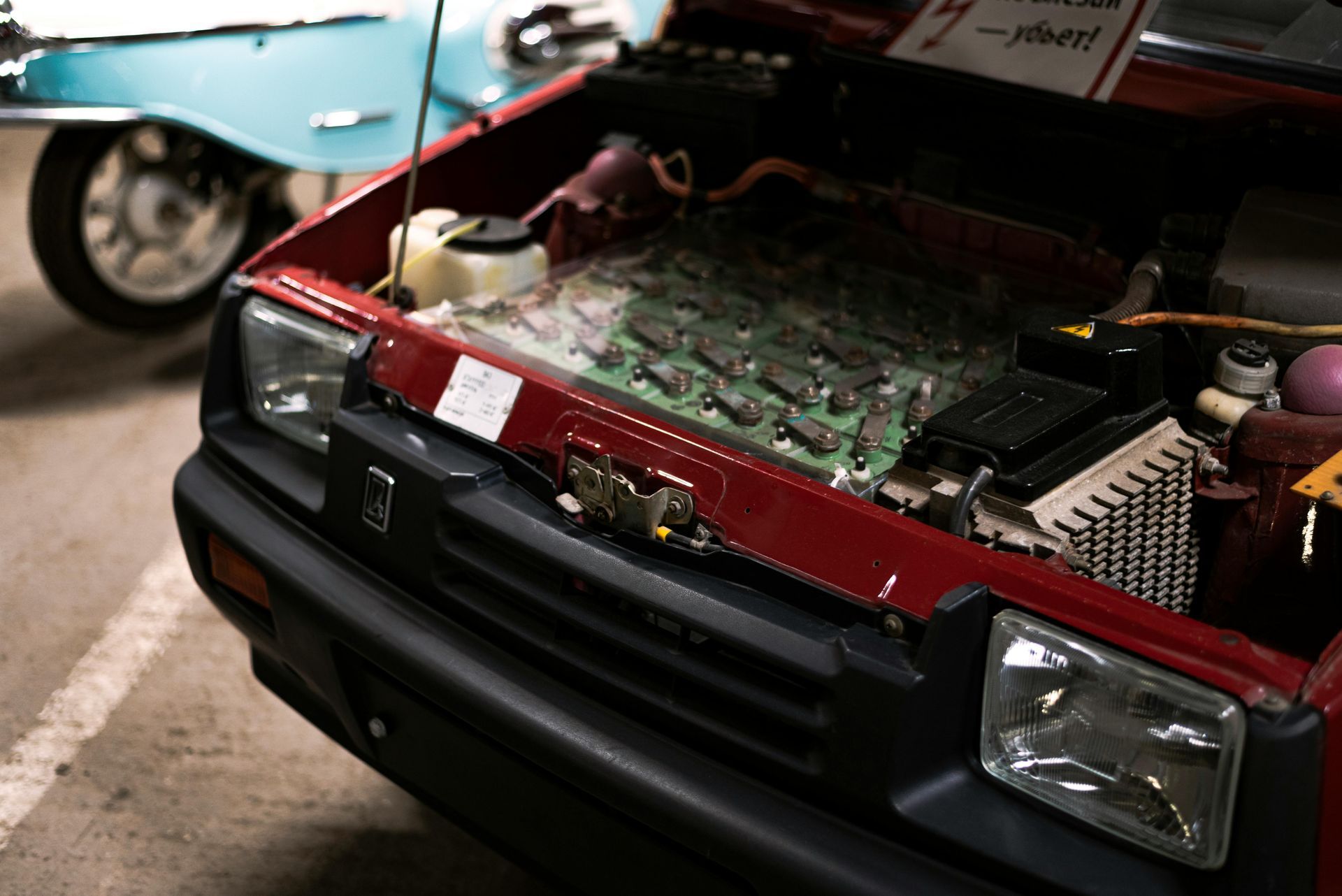 Red car with hood open, showing a computer circuit board where the engine would be, blue car in background.