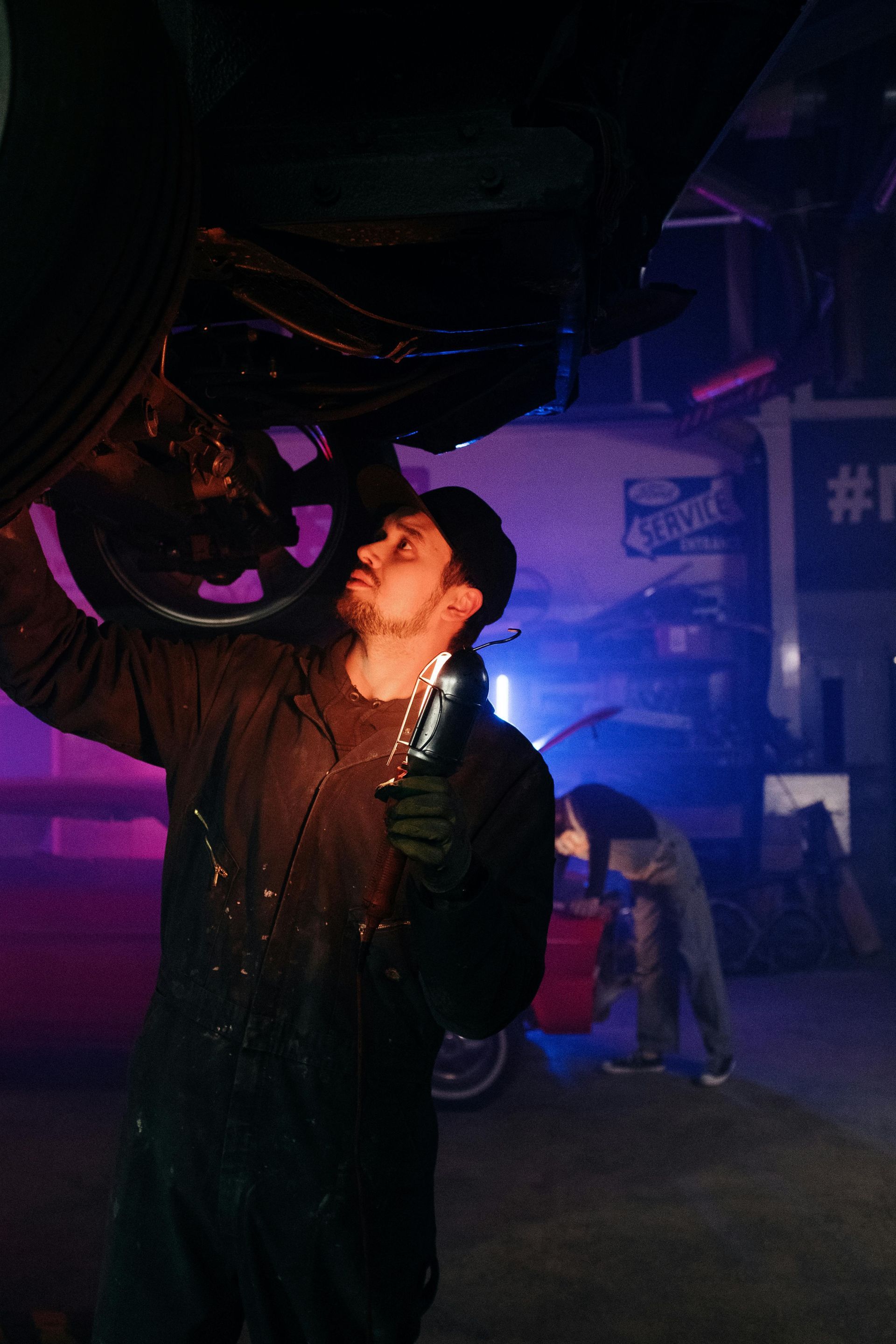 Mechanic works under a car, holding a light, in a dimly lit garage. Another person works in background.