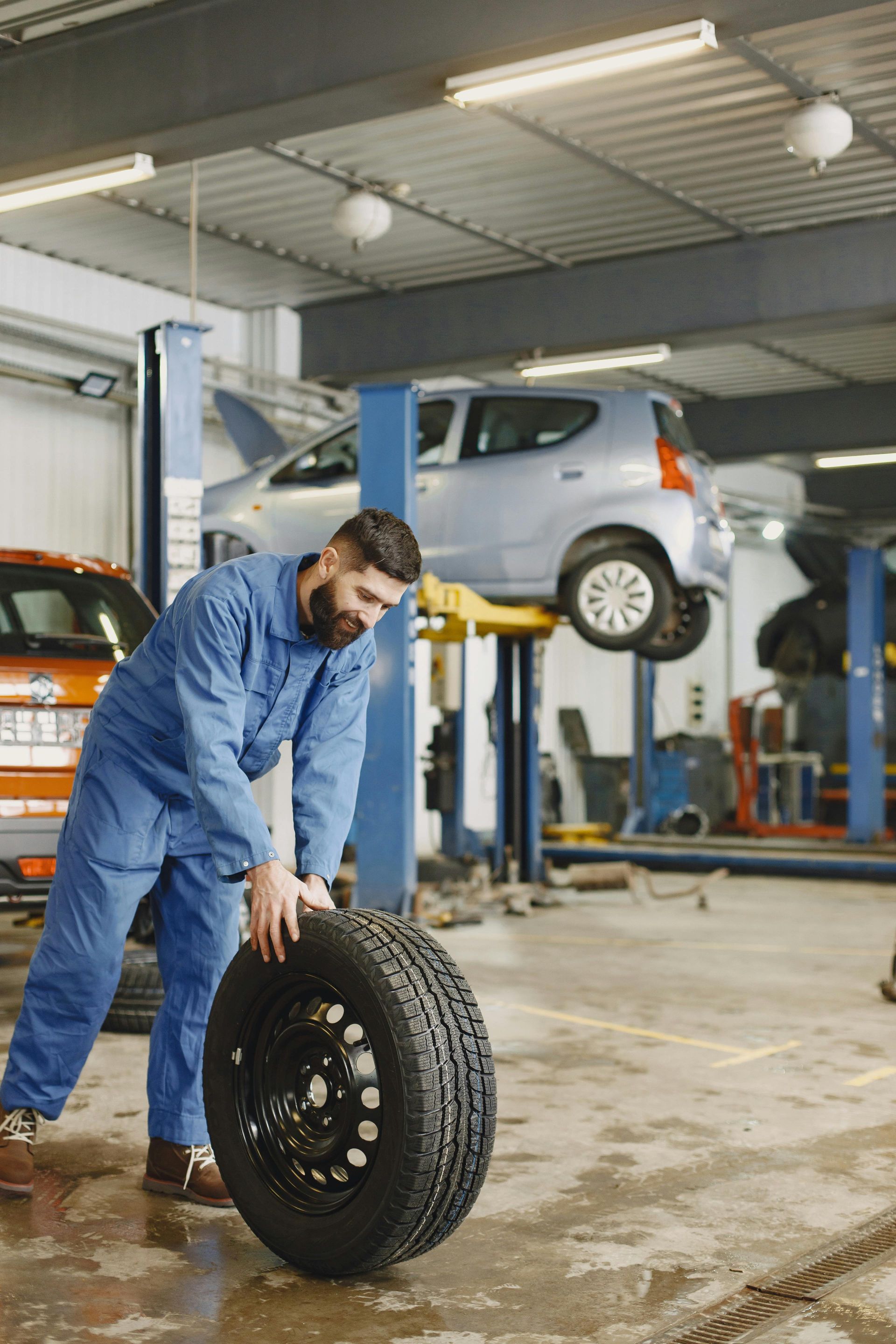 Mechanic in blue jumpsuit rolling a tire in a garage, another car raised on a lift in the background.